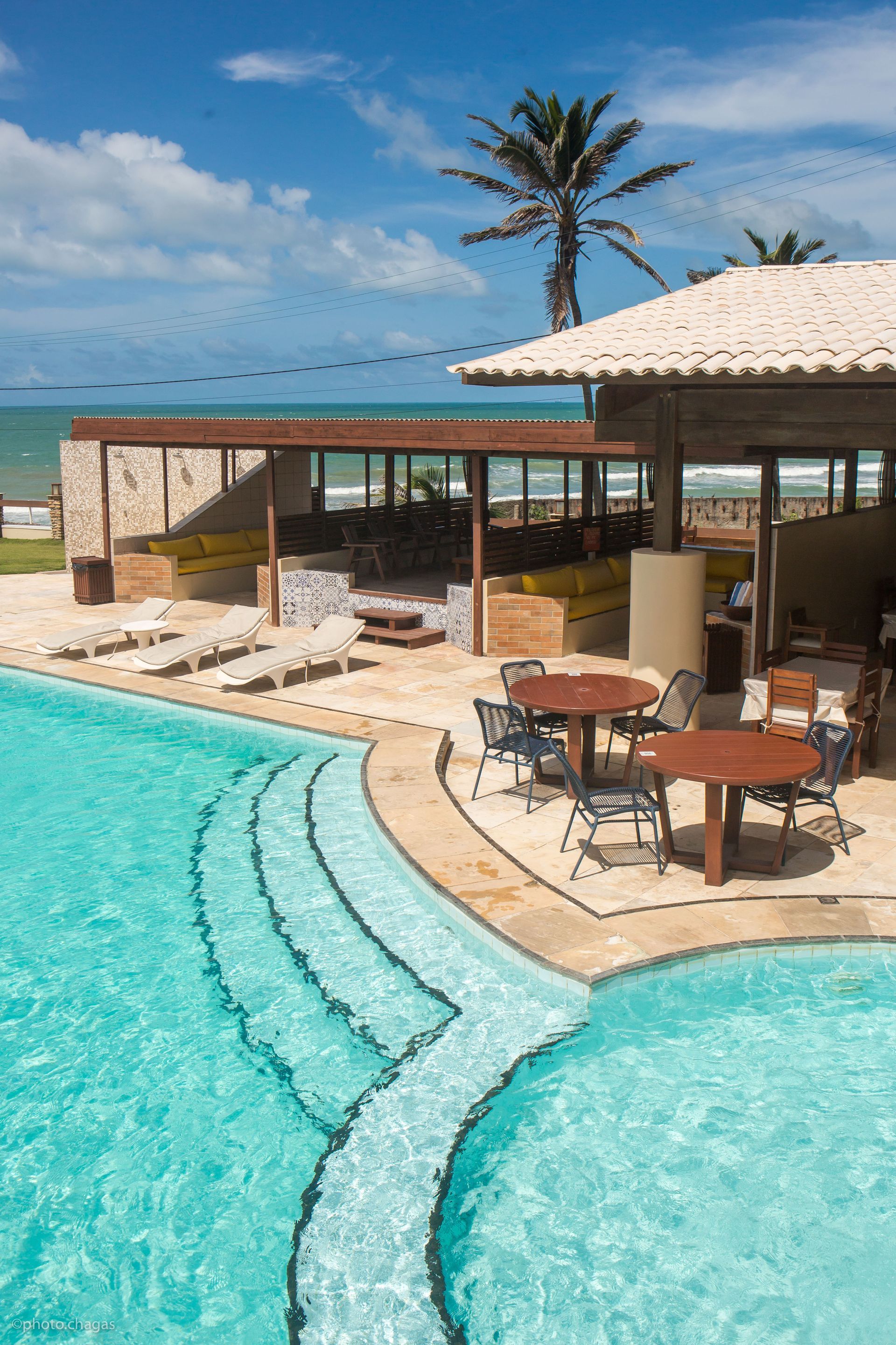 Poolside restaurant with turquoise pool, ocean view, tables, chairs, and palm tree under blue sky.