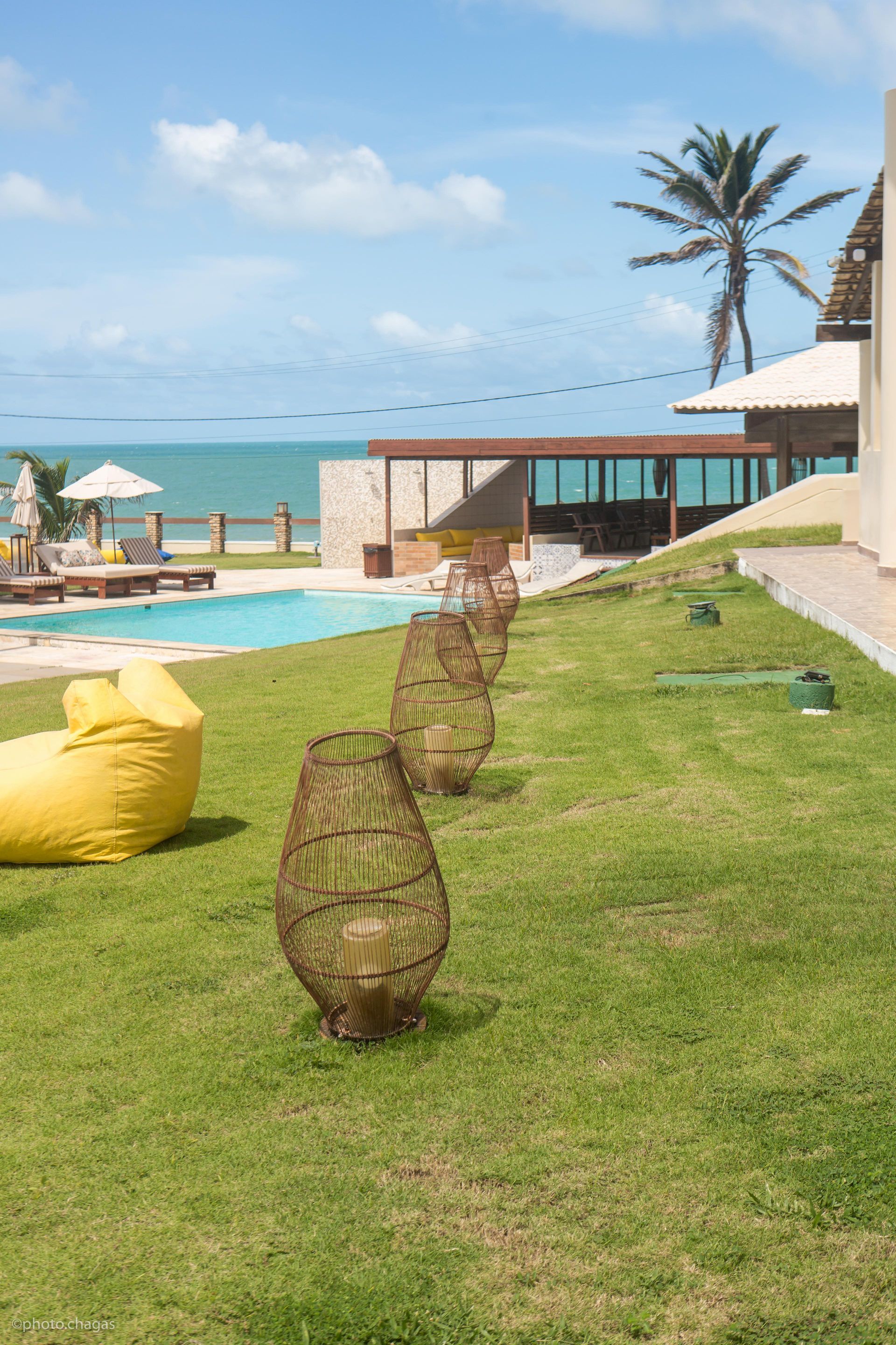 Lawn with woven lanterns leads to a pool, beach, and blue ocean under a partly cloudy sky.