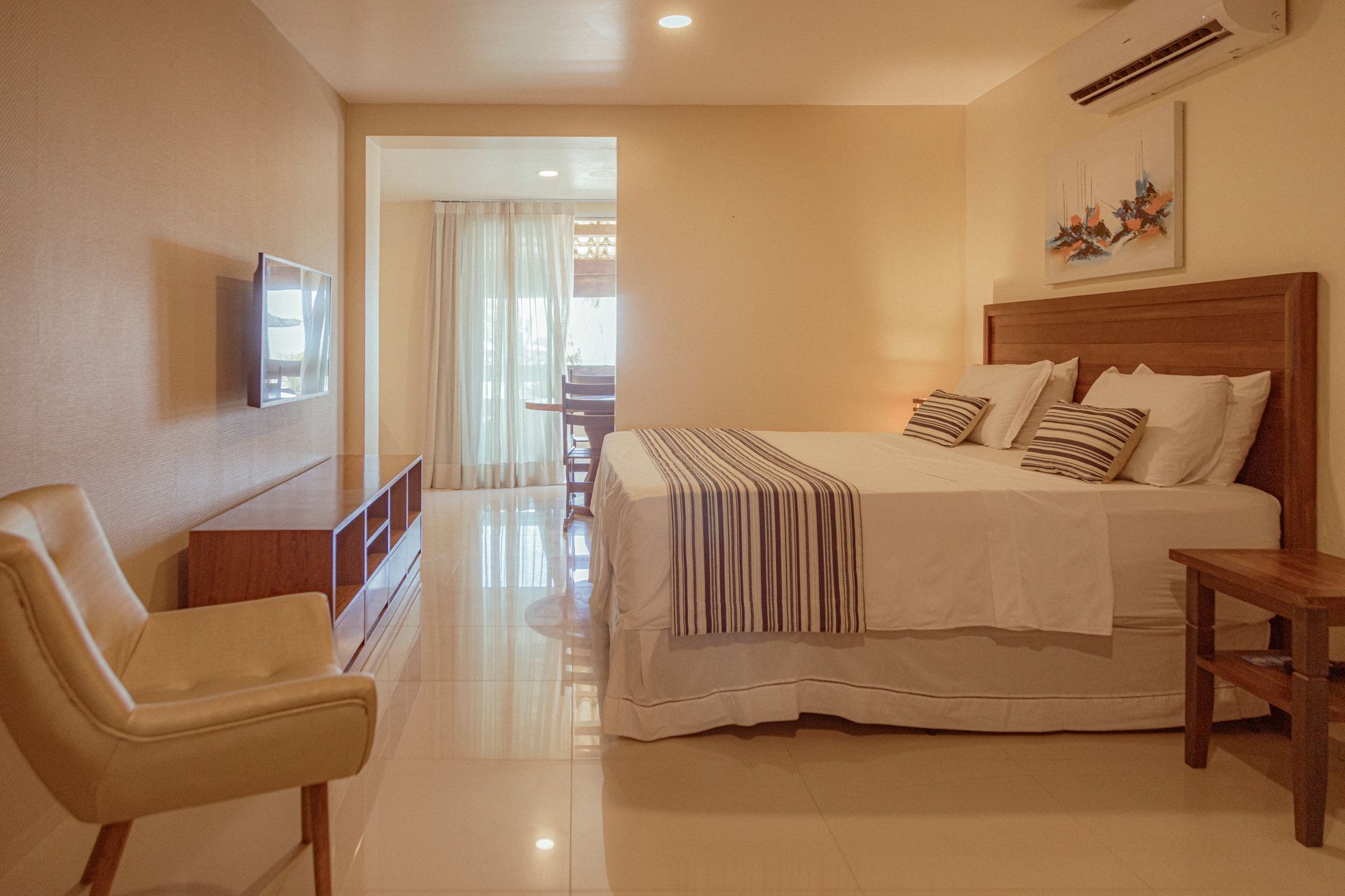 Bright, modern hotel room with bed, chair, TV, and open doorway to dining area.