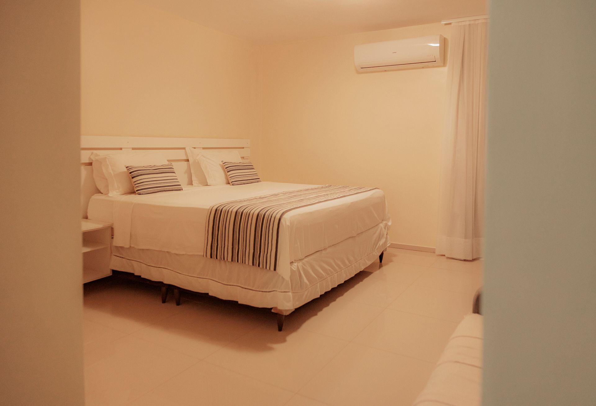 Bedroom with white bed, beige walls, air conditioner, and white curtains.