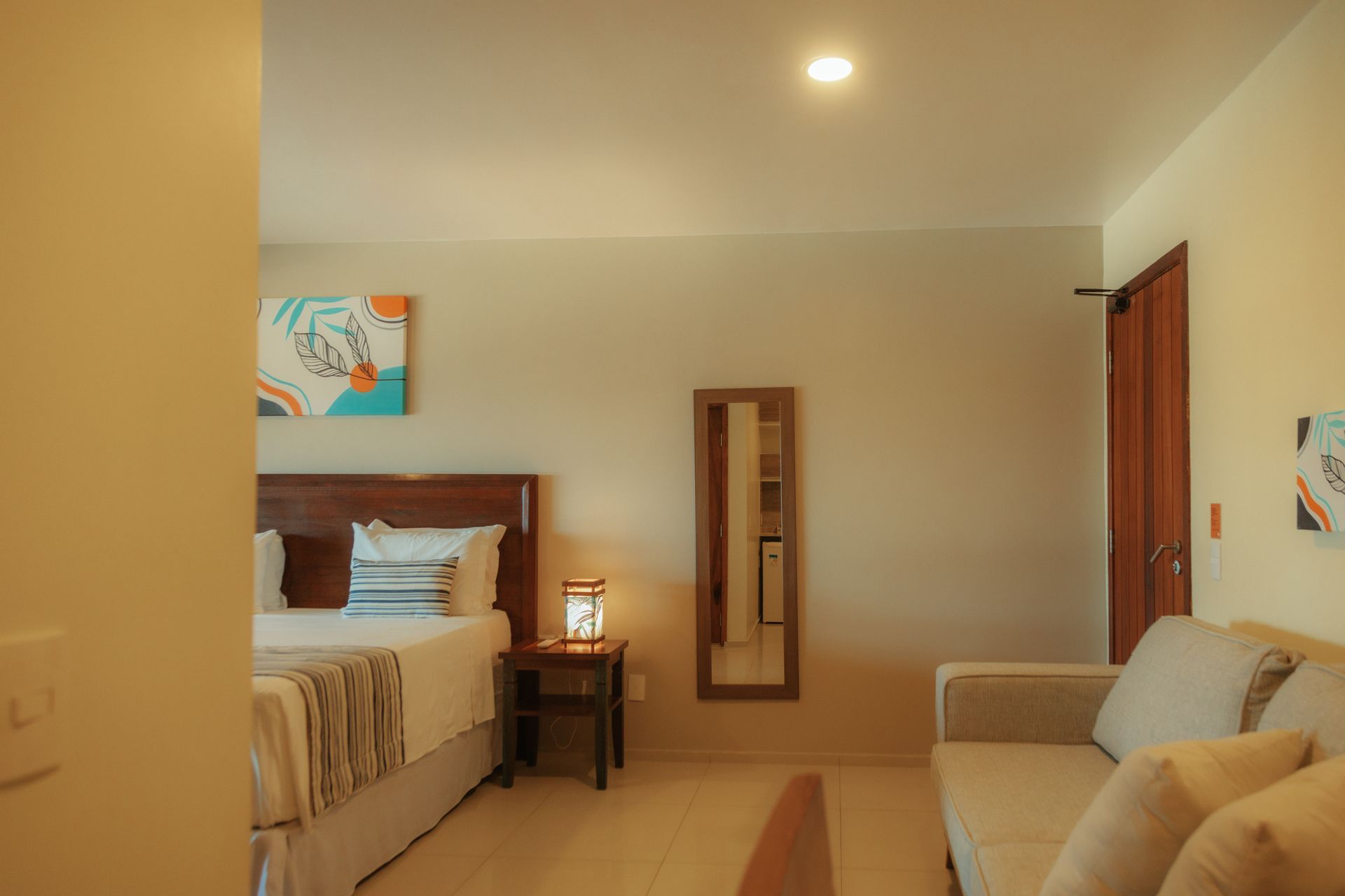 Hotel room with a bed, couch, mirror, and artwork on the wall. Light color palette.