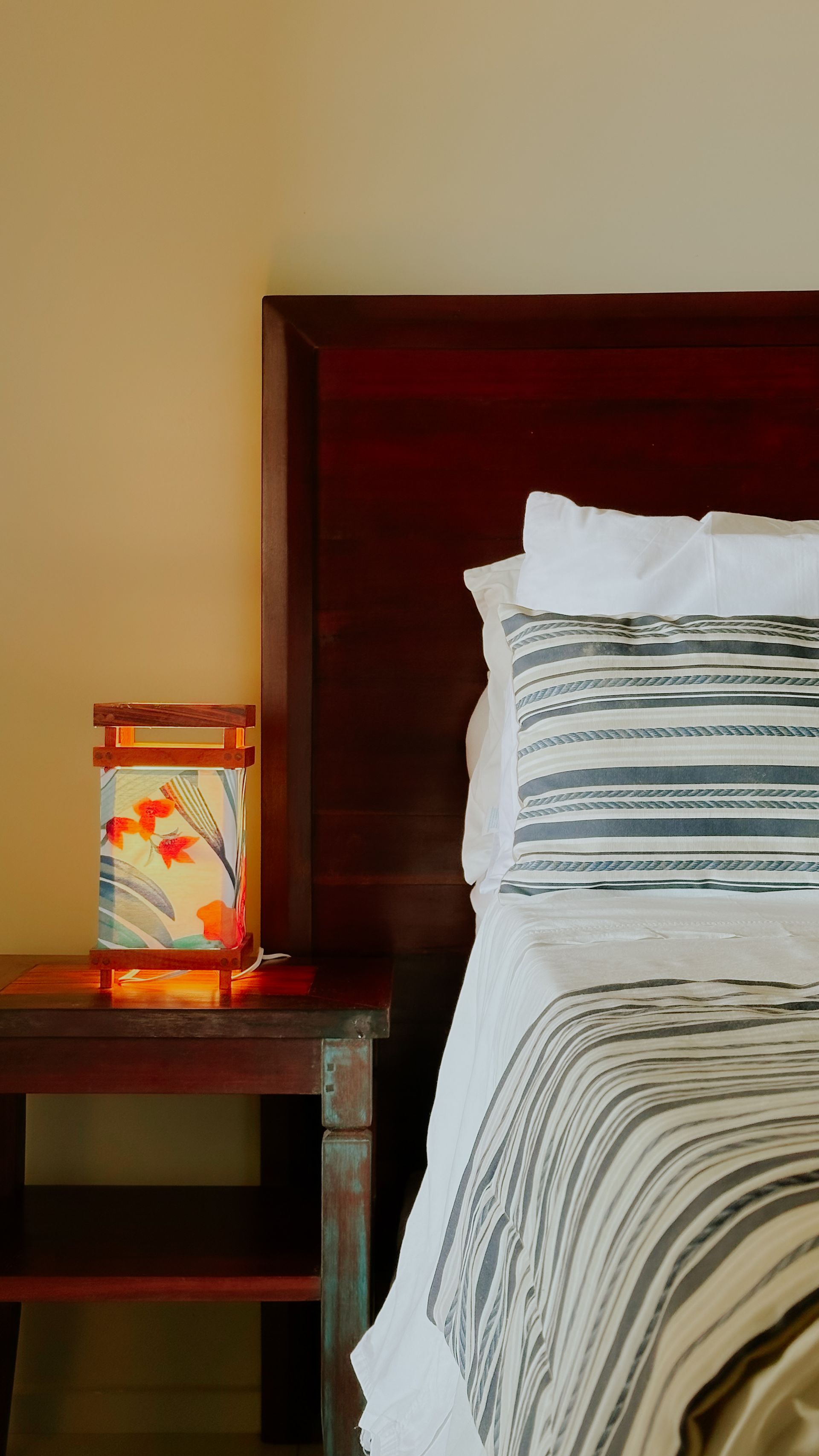 Bedside table with lamp, bed, and headboard. The lamp is rectangular with a floral design.