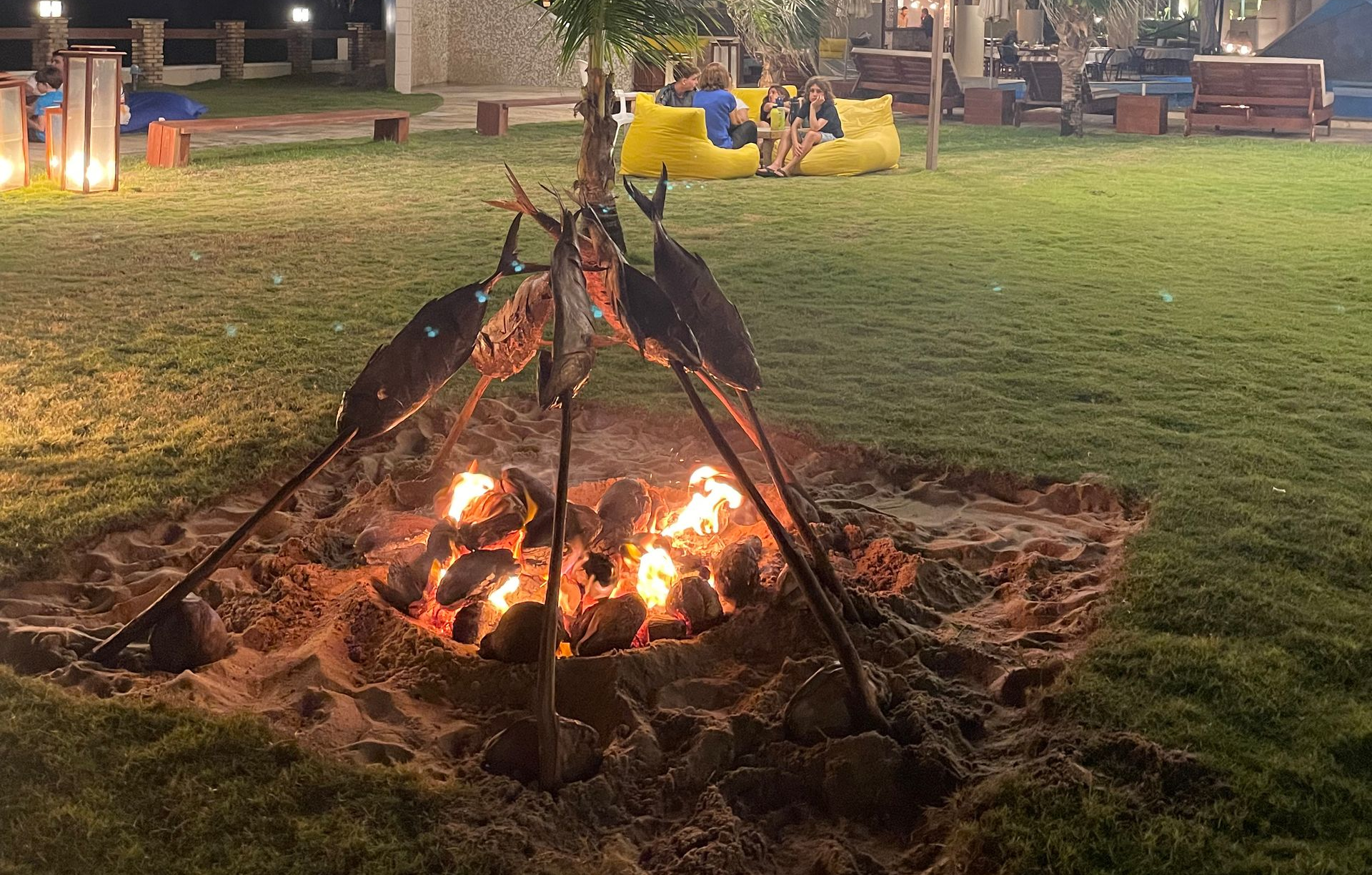 A small fire in a sandy pit, framed by metal supports. People sit nearby on yellow bean bags in a grassy area at night.