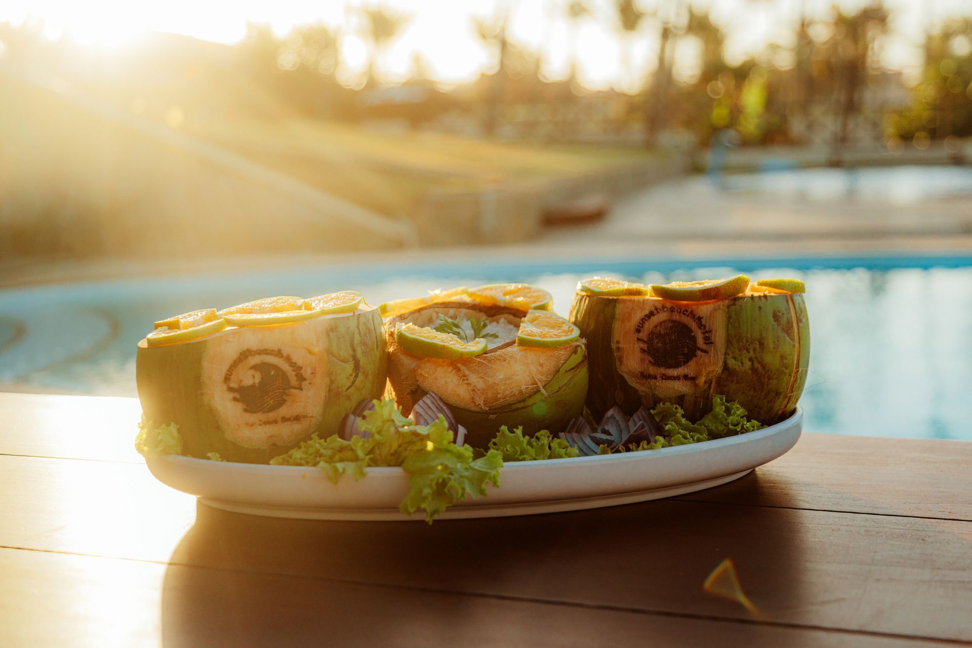 A platter with three halved coconuts filled with food, poolside at sunset.
