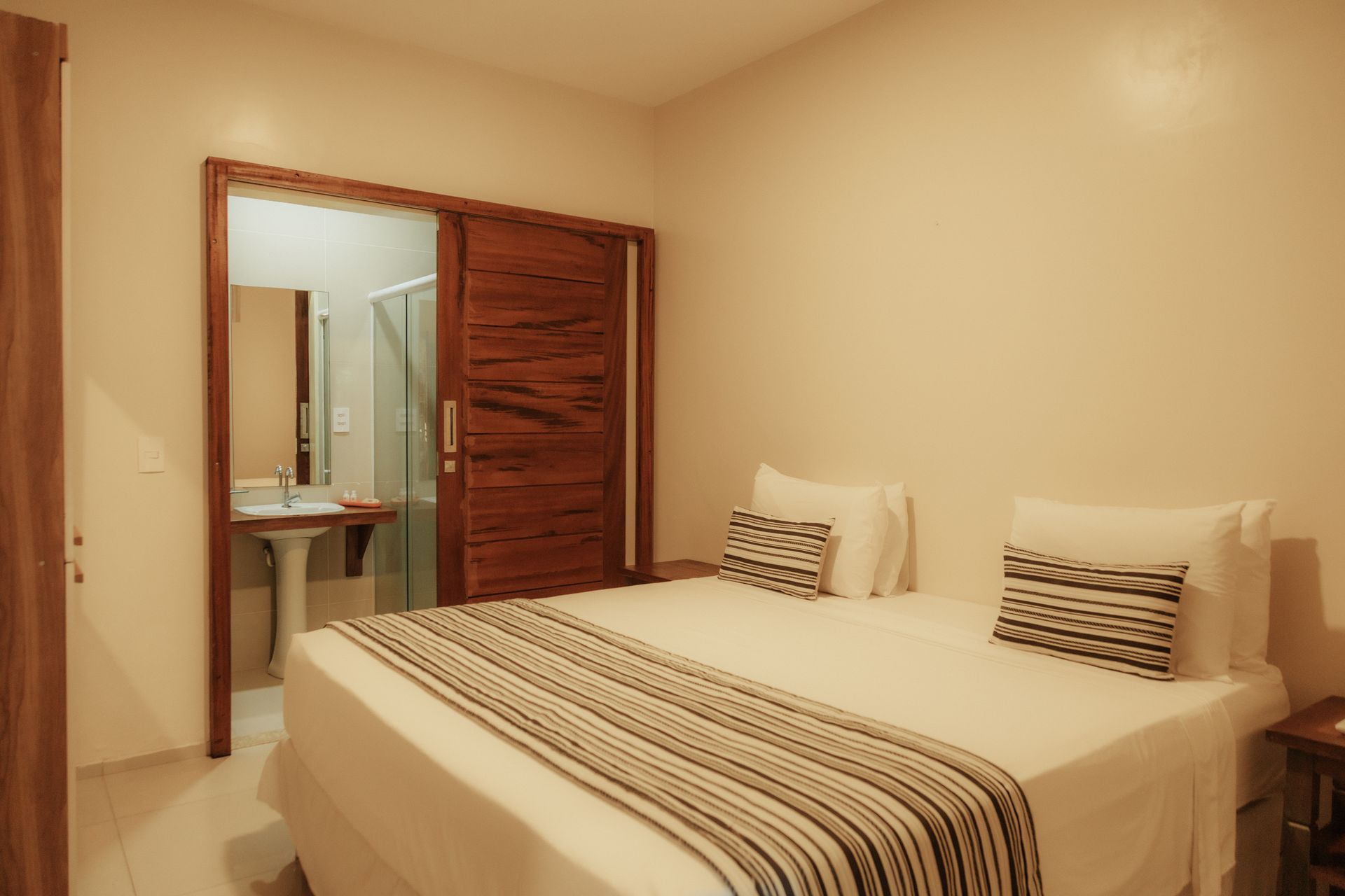 Bedroom with a double bed, open wooden door to a bathroom with sink, white walls, warm lighting.