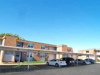 A row of cars are parked under a covered parking lot in front of a brick apartment building.