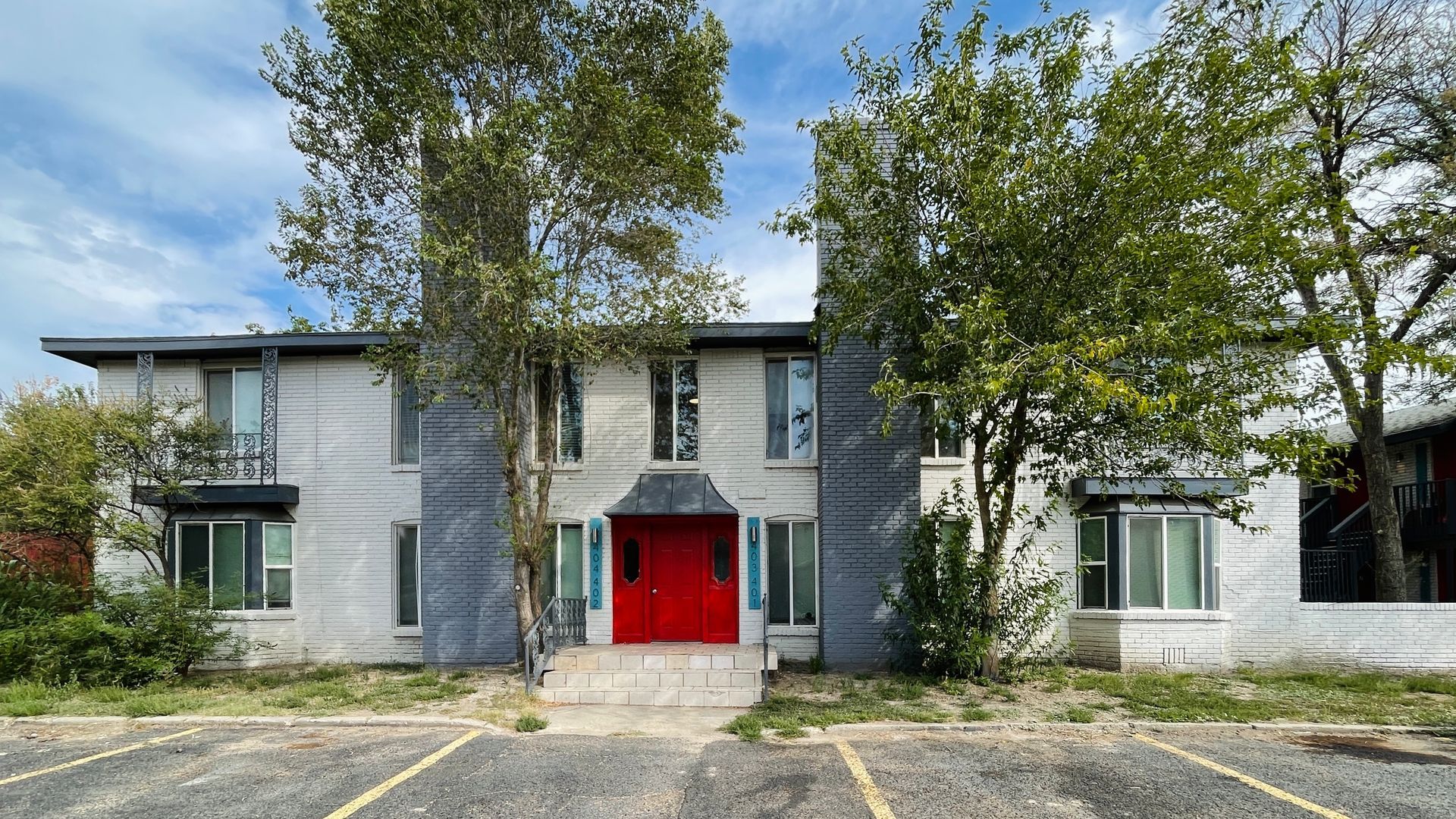 A white apartment building with a red door and a parking lot in front of it.
