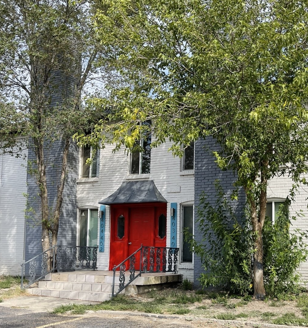 A white house with a red door and blue windows is surrounded by trees.