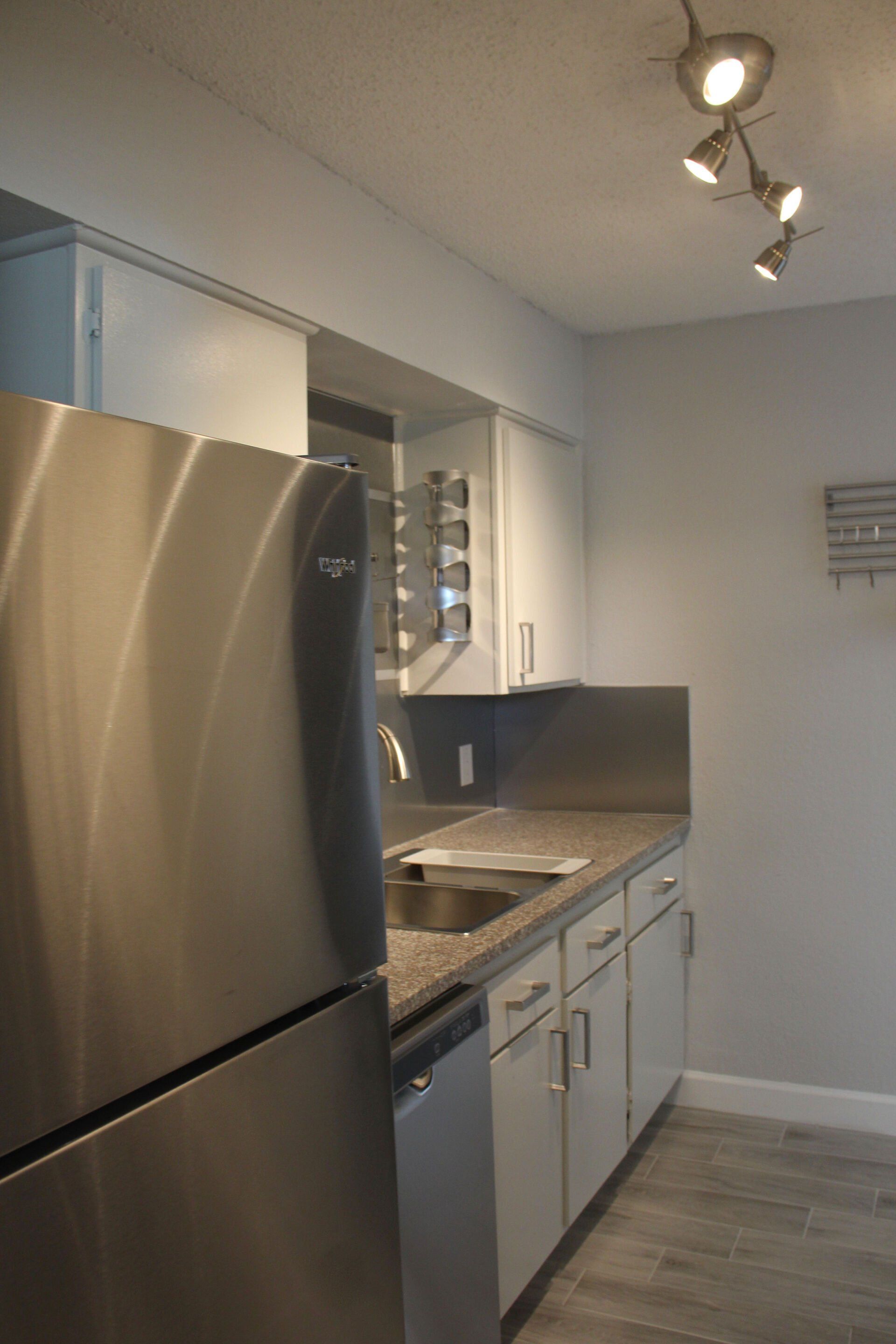 A kitchen with a stainless steel refrigerator , sink , and dishwasher. Granite countertop & stainless steel backsplash & wine racks