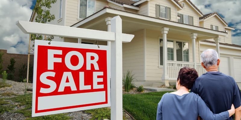 Seniors standing in front of a home with a For Sale sign in front of it