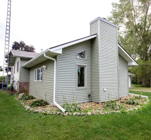 Green-sided house with a large chimney, white trim, and a small garden, set on green grass.