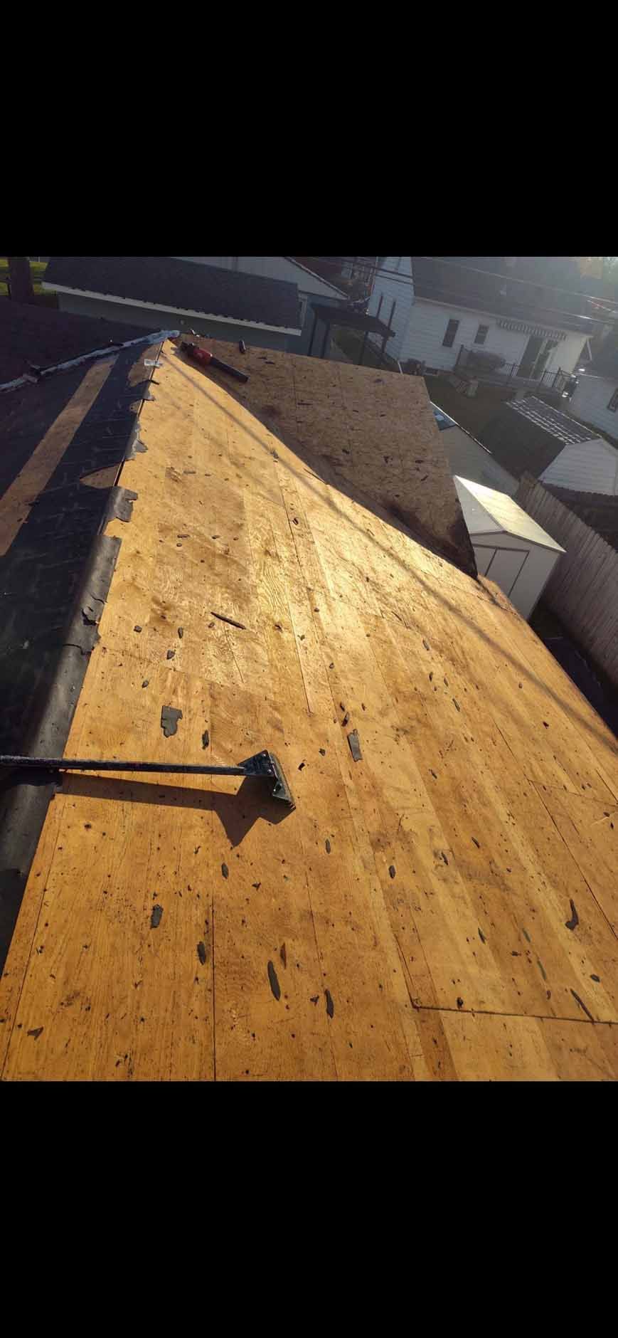 View of a roof with exposed plywood boards. A tool rests on the roof. Buildings are visible in the background.