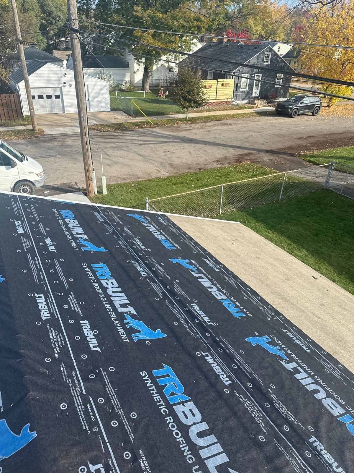 Roof under construction, showing black synthetic roofing underlayment, residential neighborhood in background.