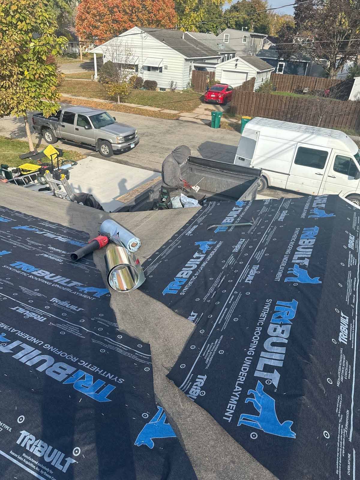 Man working on a roof, applying roofing material. Suburban setting with truck and van.