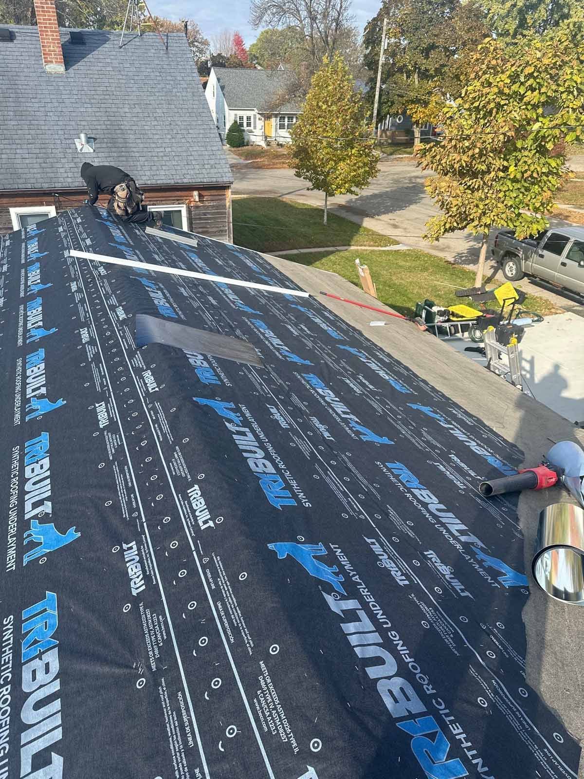 Roofer on a house roof installing underlayment. Sunlight, a street, trees, and other houses are in the background.