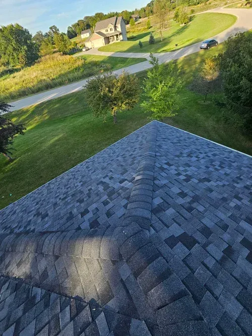 Dark gray asphalt shingle roof, green lawn, trees, road, and house in background.