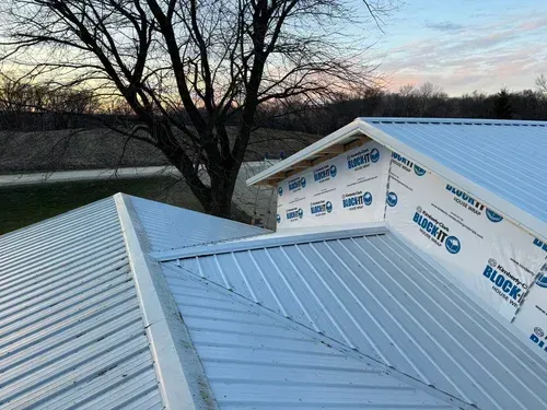 A metal roof on a building with white siding, with trees and sky in the background.