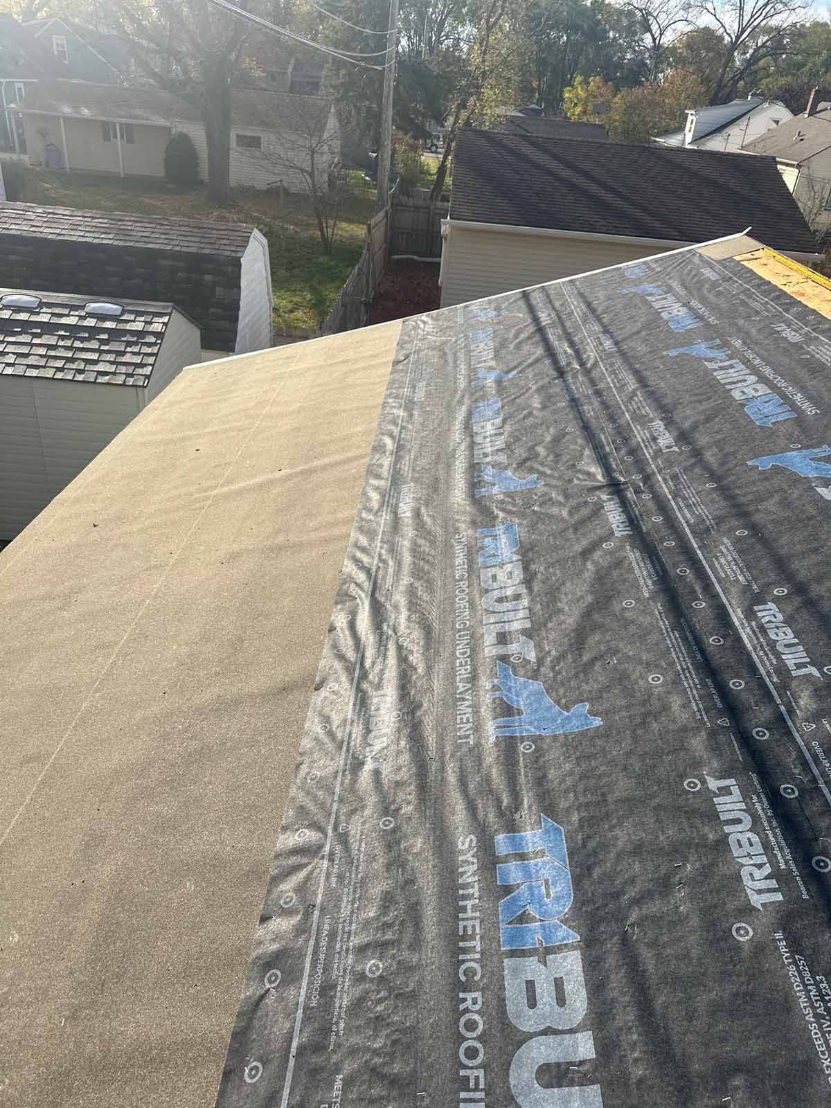 Rooftop with black synthetic underlayment partially covering brown roof. Trees and houses in background.