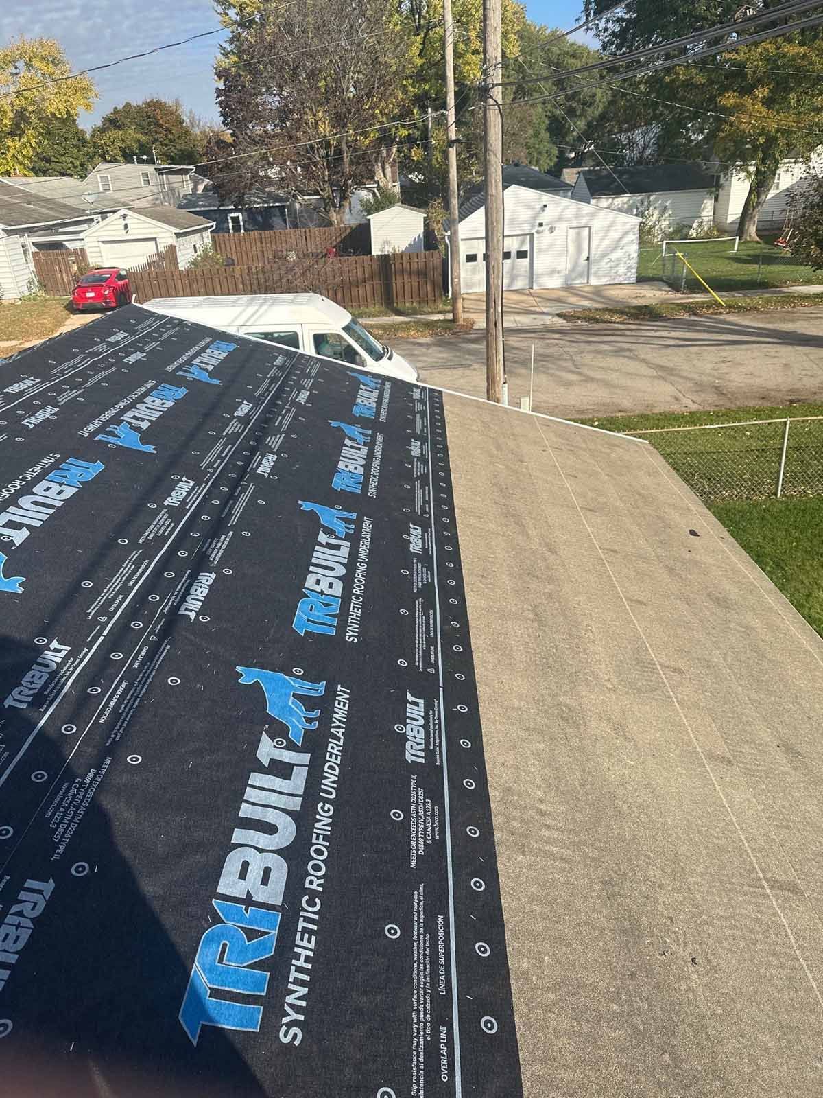 Close-up of a roof edge with synthetic roofing underlayment being installed. Houses and a street are in the background.