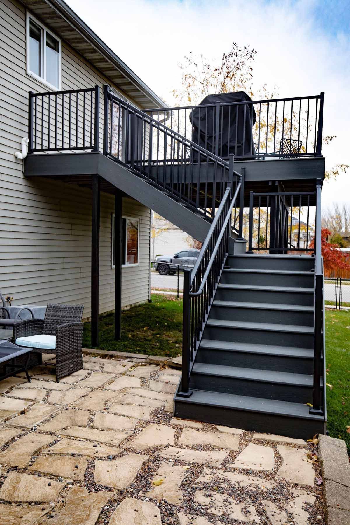 Two-story outdoor deck with black railing and steps; a barbecue grill sits on the upper level, next to a house.