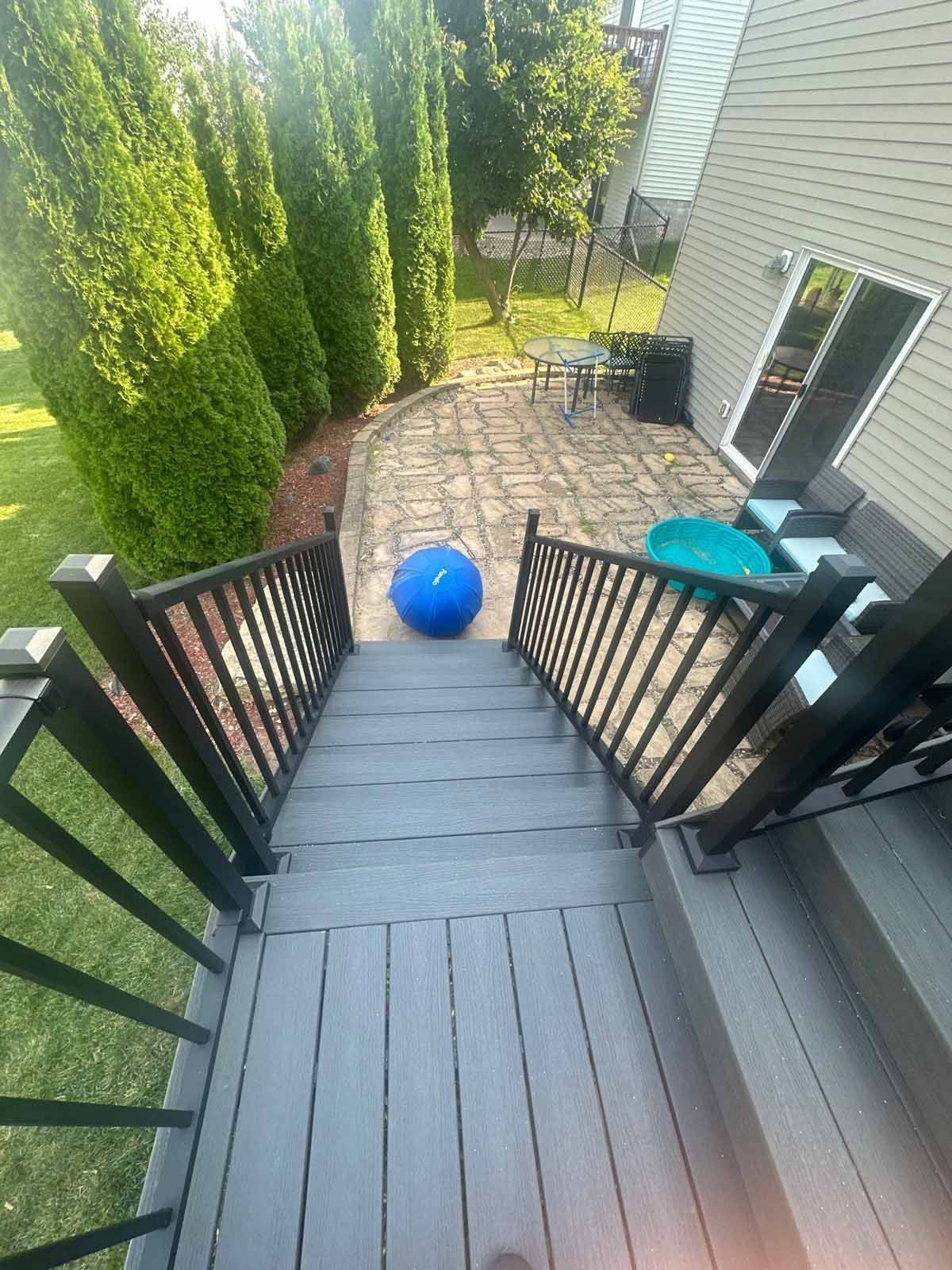 Deck stairs leading to a patio with a large blue ball, green chairs, and a table surrounded by trees.