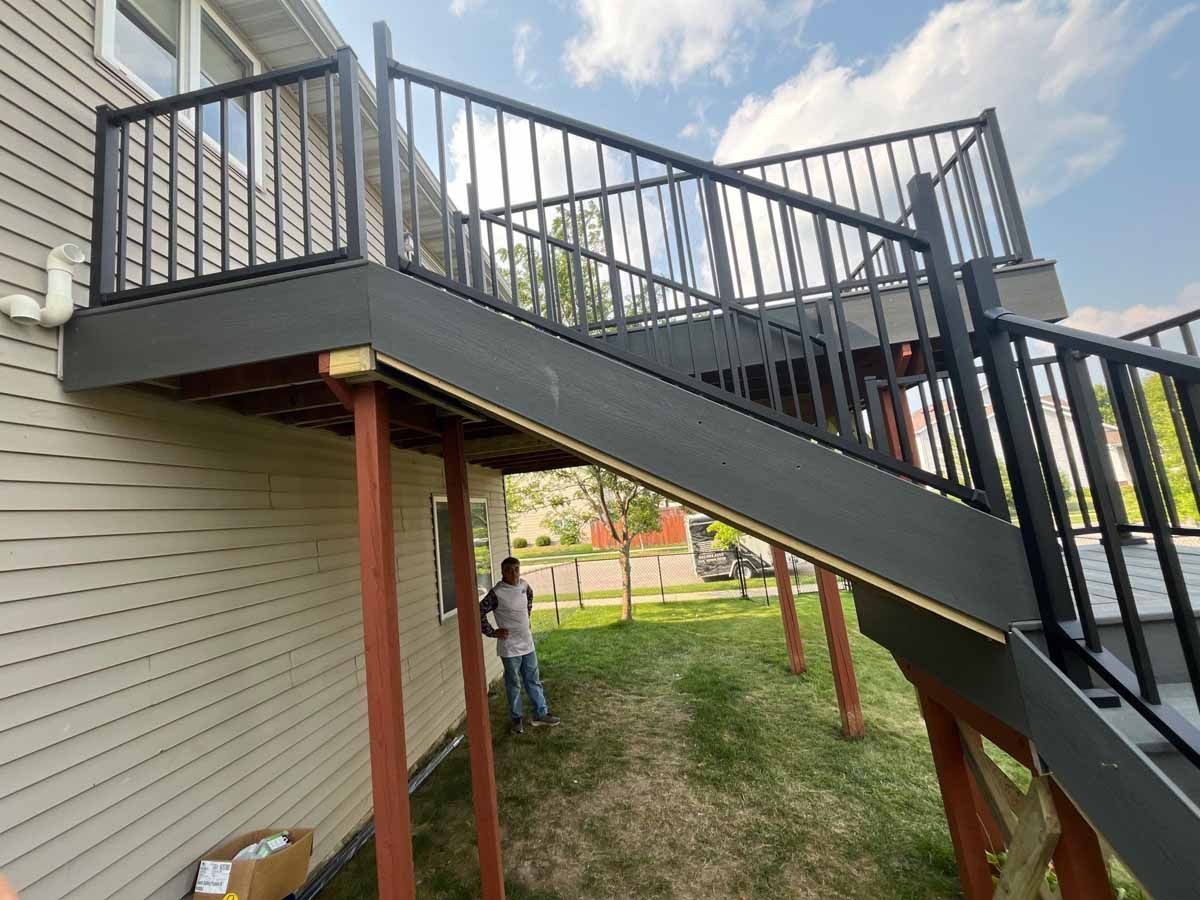 Dark gray deck with black railings and steps. A person stands beneath, near a light-colored house.