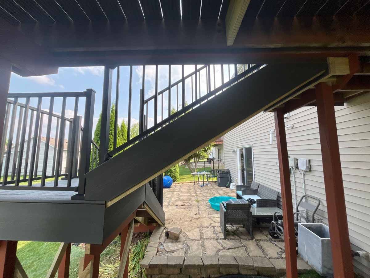 Staircase leading down from a deck, black railings, gray stairs, wooden supports, backyard view.