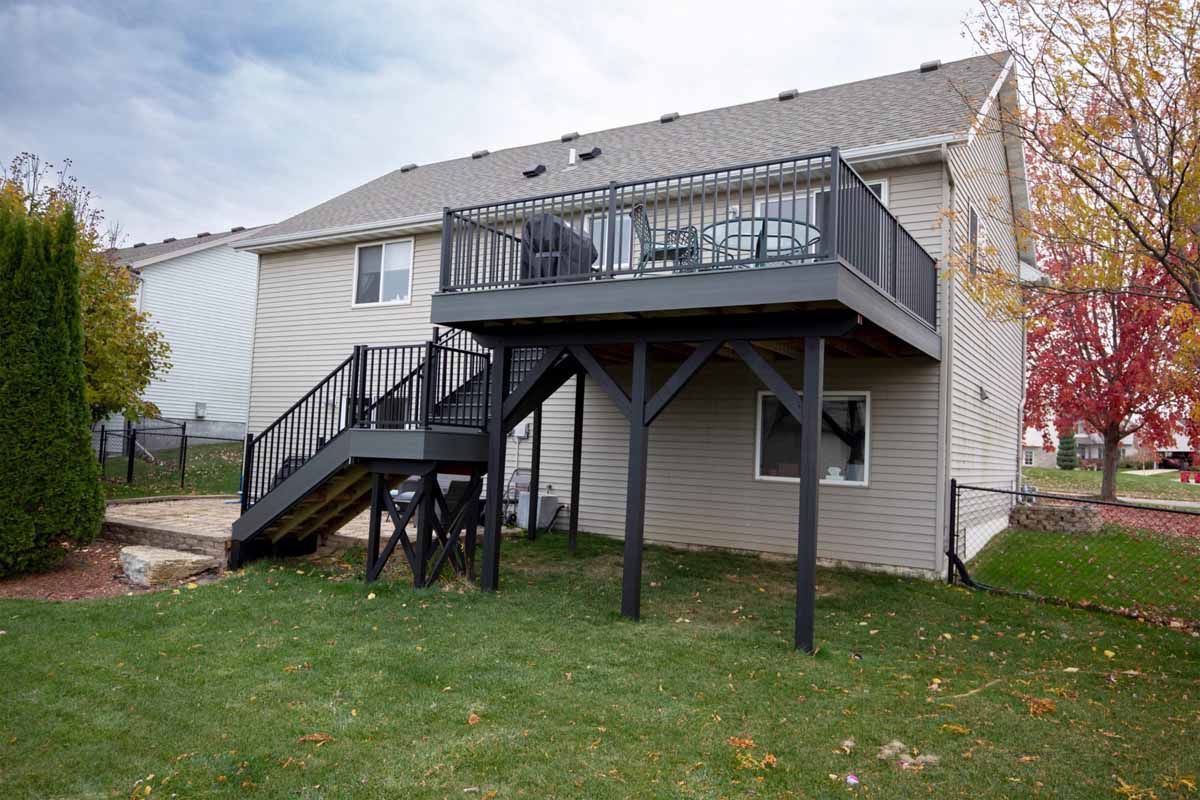Two-story house with a grey deck. Black railing, stairs, and supports. Green lawn and fall foliage.