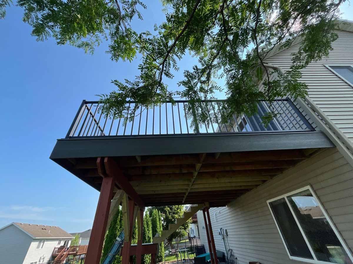 Low-angle view of a deck with black railing against a blue sky, beneath a tree. Wooden deck support.