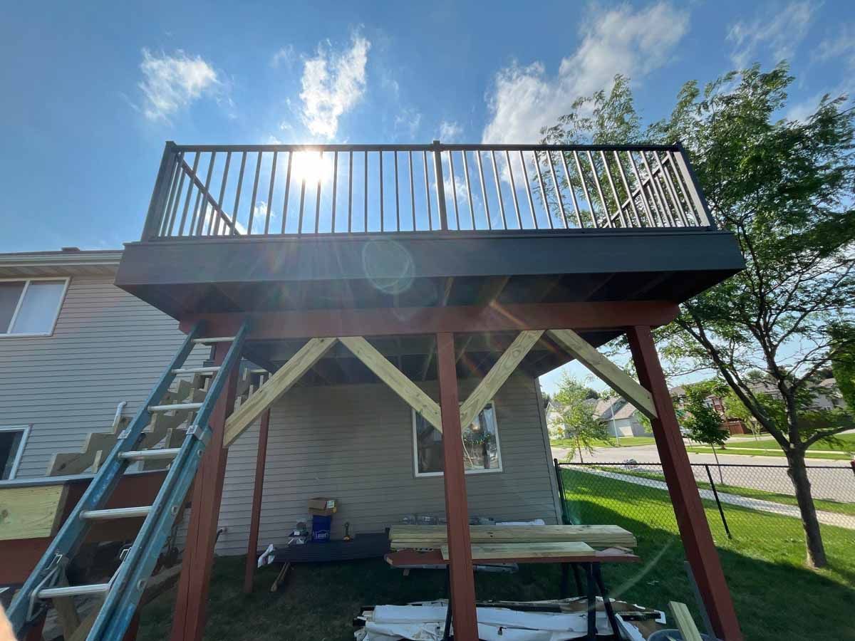 Elevated wooden deck attached to a house with railing. Supports and ladder visible. Bright sunny day.