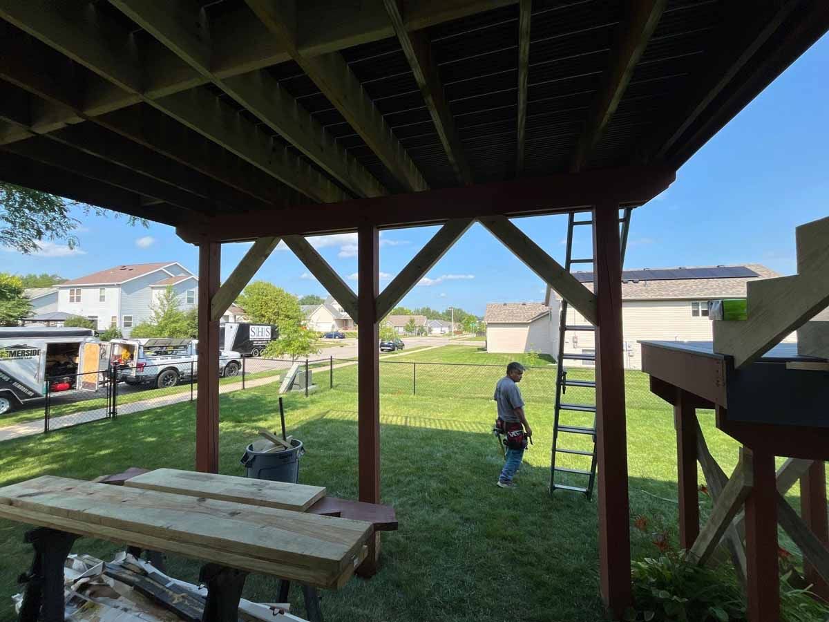 Under-deck view; man on ladder in backyard, wooden posts, new deck boards. Green grass, blue sky, houses in the distance.
