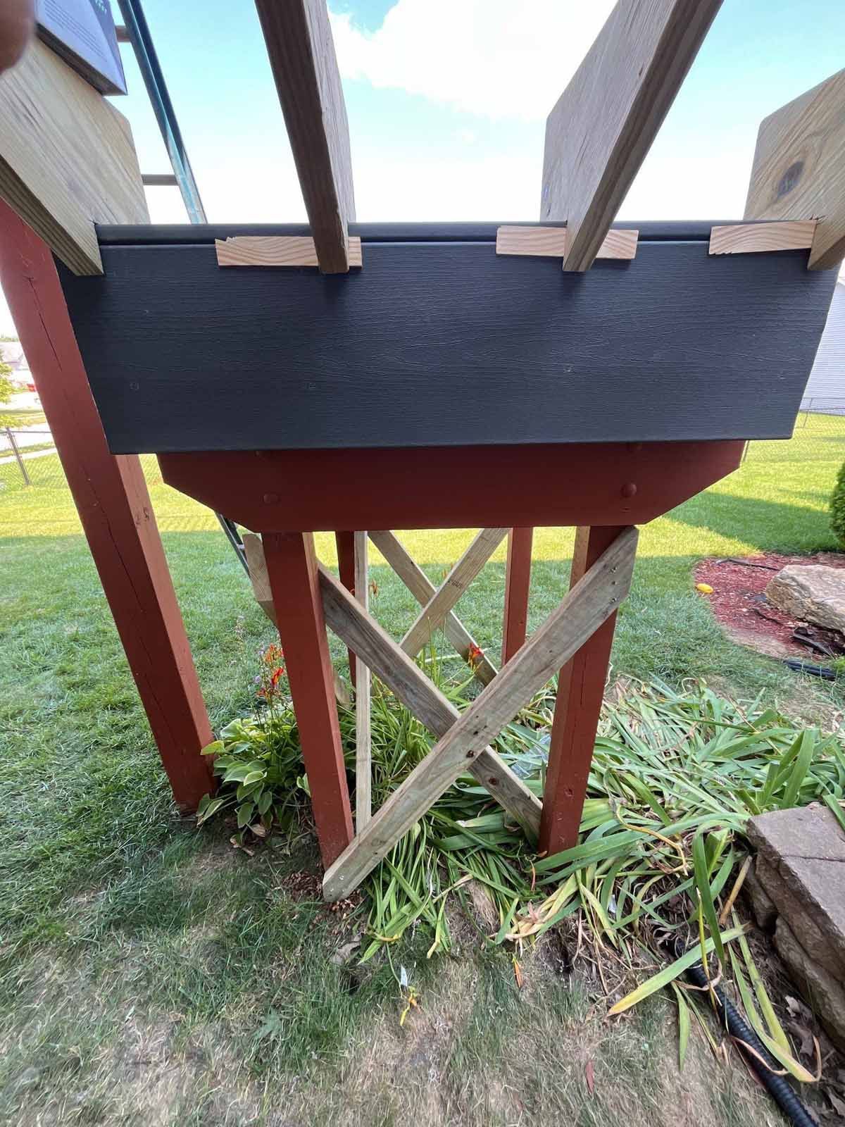 Wooden structure with black top, supported by red legs with angled bracing, in a grassy yard.