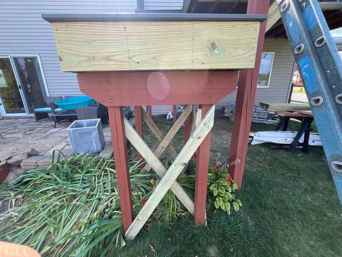 Wooden planter box on a raised platform supported by red-painted posts, green X-braces, and green grass.