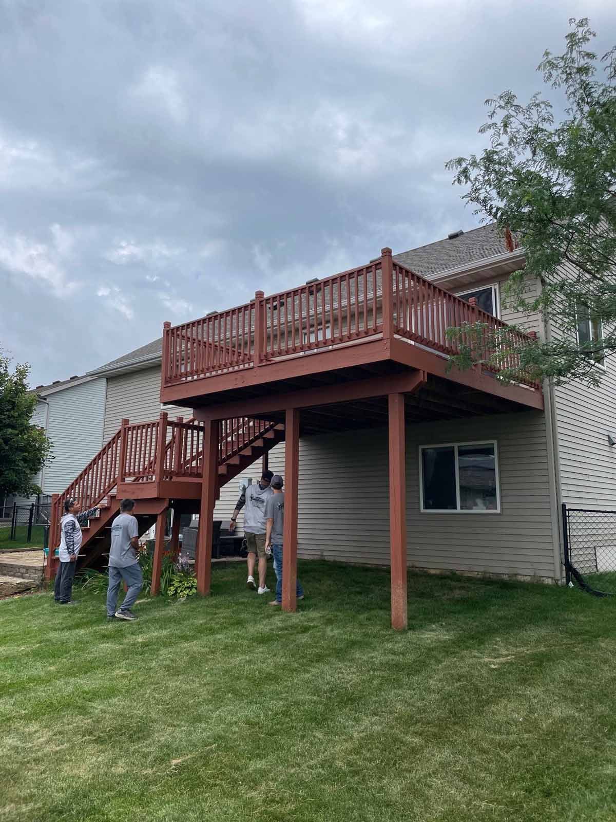 Wooden deck attached to a house with stairs. People stand on the lawn below. Overcast sky.