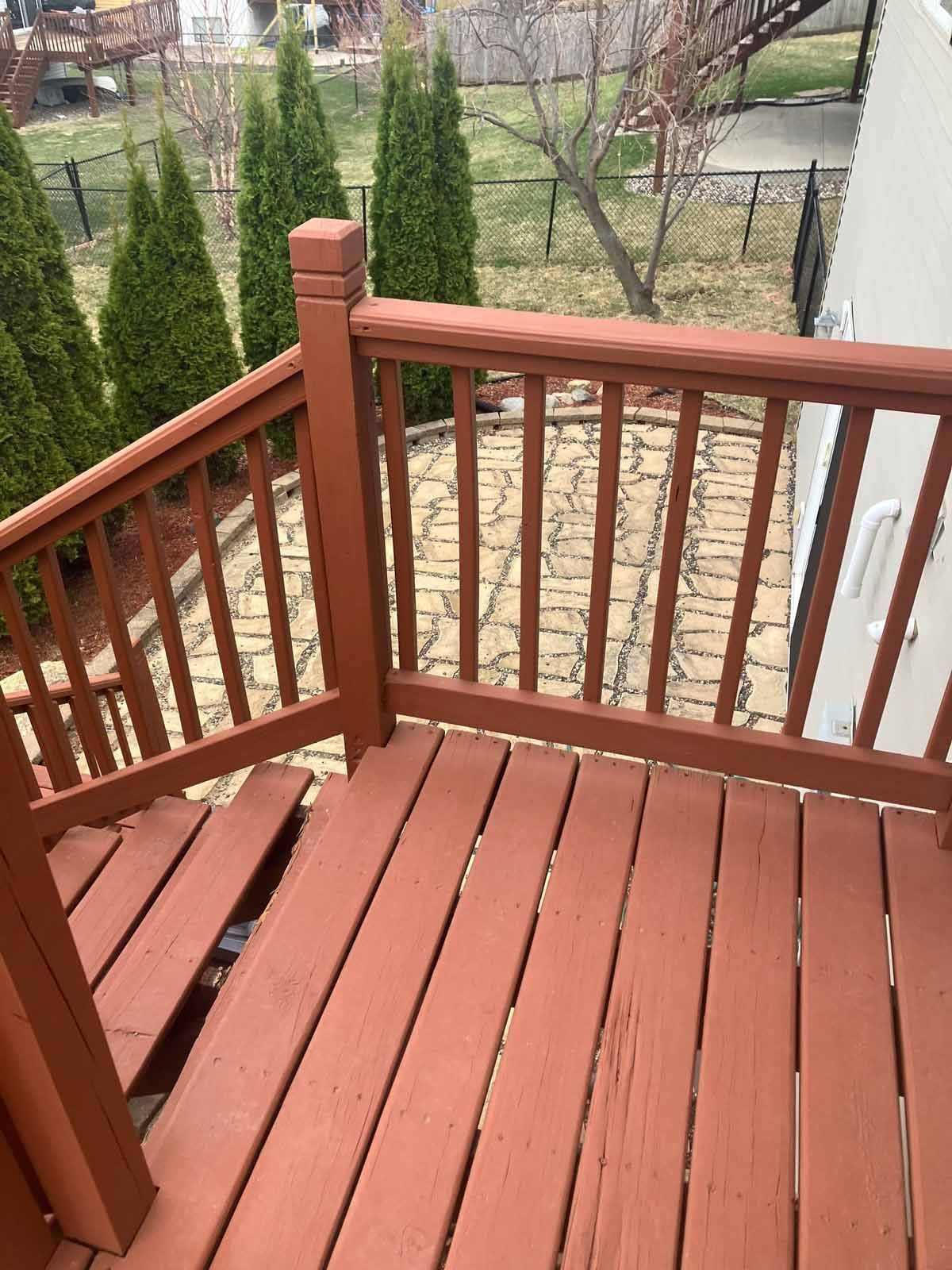 Brown wooden deck with railings, overlooking a paved patio area with green trees in the background.