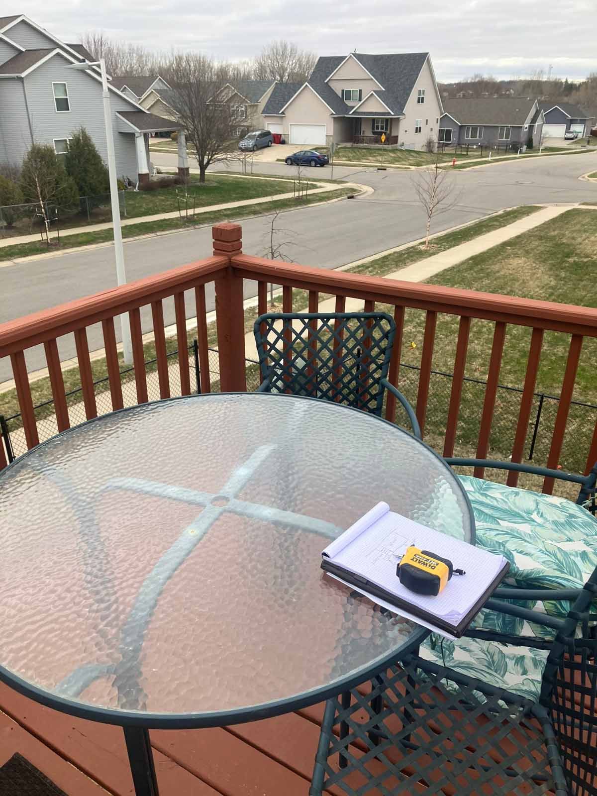 A glass-top patio table with notebook and tape measure, surrounded by chairs on a wooden deck overlooking a suburban street.