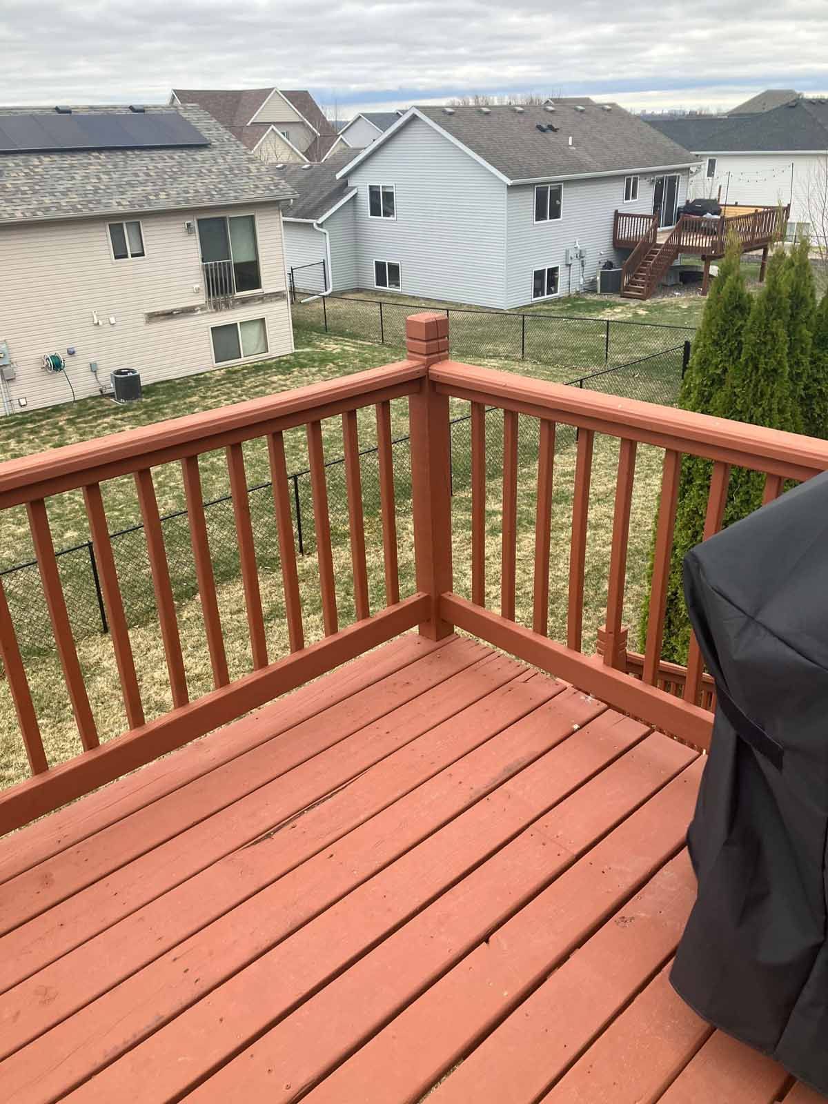Red wooden deck with railing, overlooking grassy yards and houses under a cloudy sky.