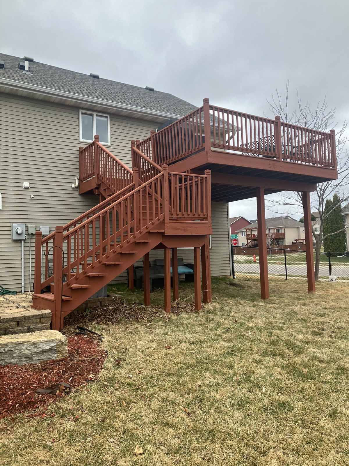 Two-story wooden deck painted brown, attached to a house with stairs. Overcast day.