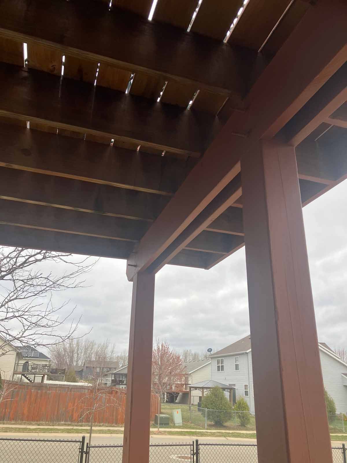 View from beneath a brown wooden porch roof with support beams, overlooking a fence and suburban houses on a cloudy day.
