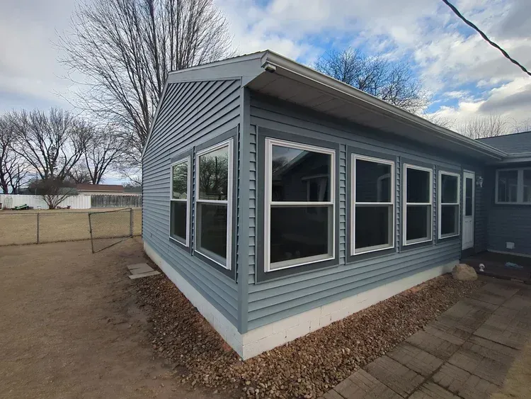 Blue-sided sunroom addition with large windows, set on a gravel base next to a house with a clear sky.