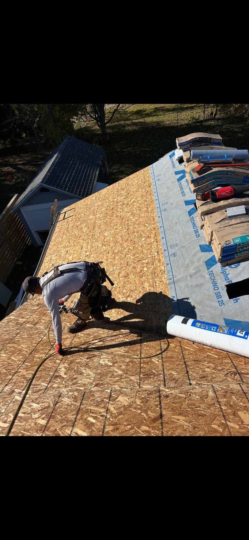 Roofer working on a partially shingled roof, holding tools and wearing safety harness. Houses and trees are in the background.