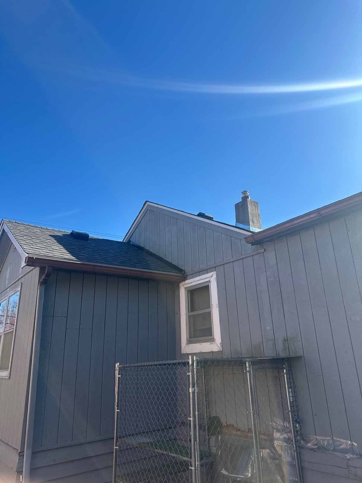 Side of a house with gray siding, a small window, and a roof, under a blue sky.