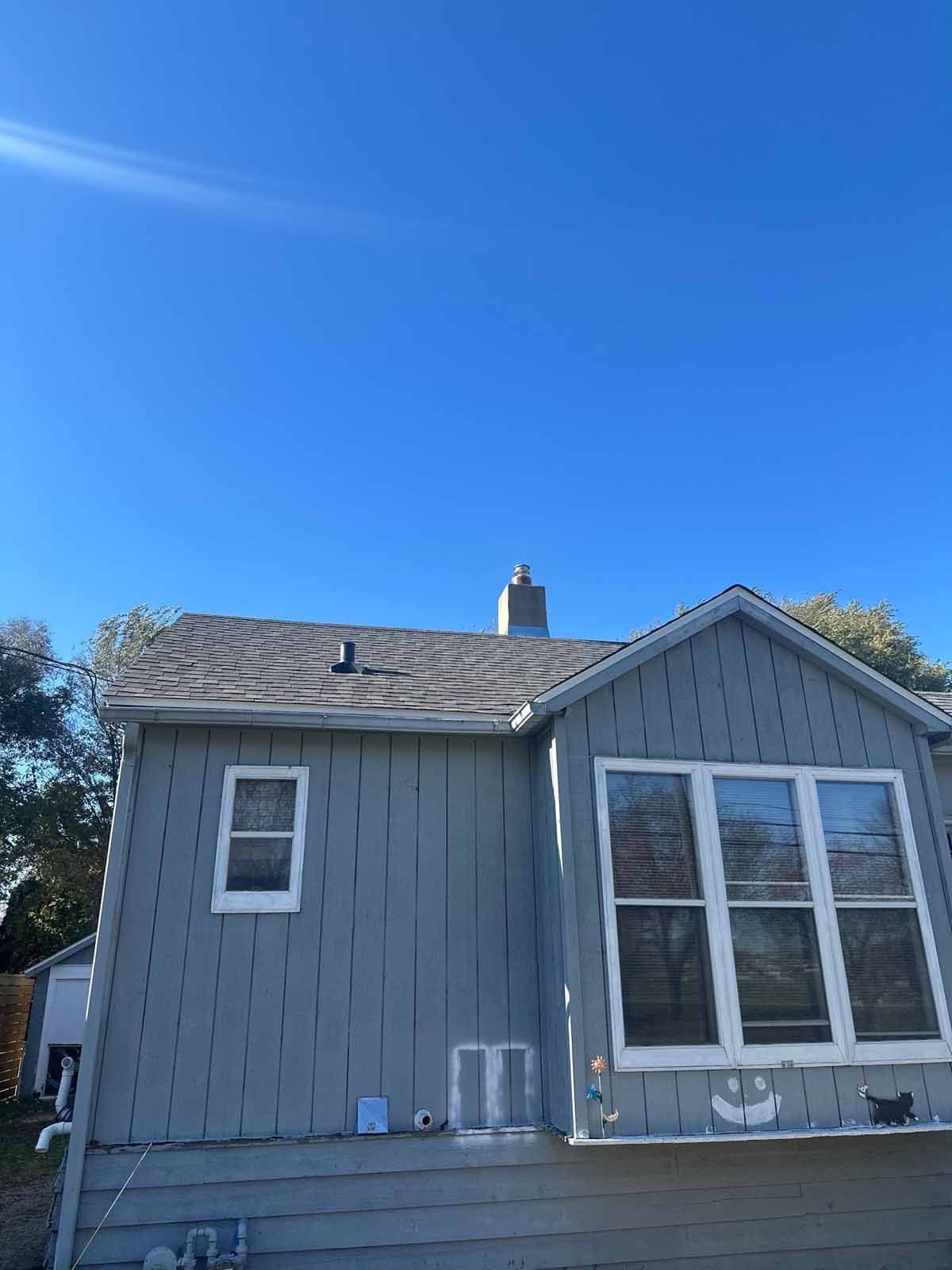 Blue-sided house with a gray roof and chimney against a clear blue sky. White window frames.