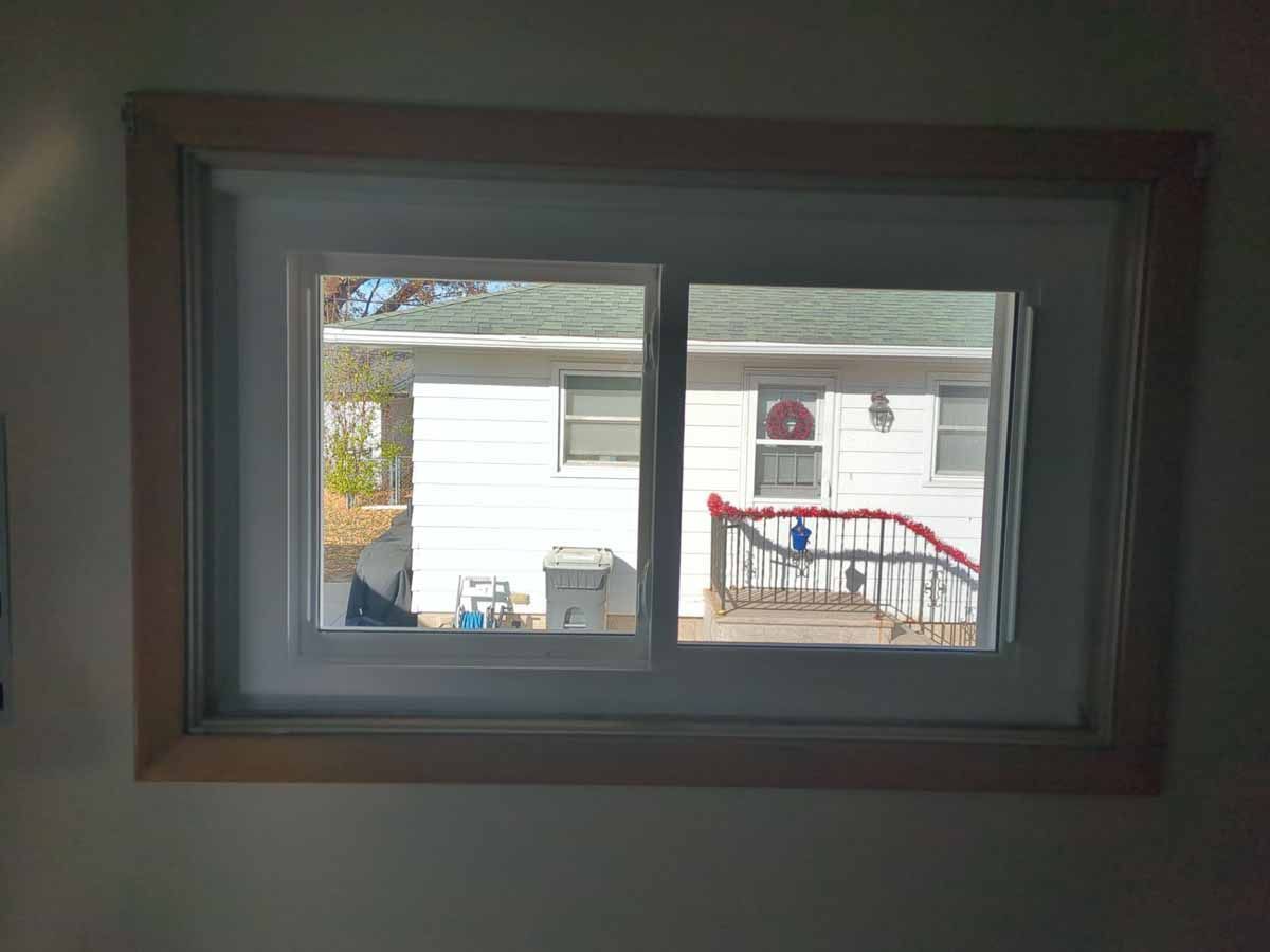 Window with a view of a white house with a red railing. A trash can and foliage are visible.