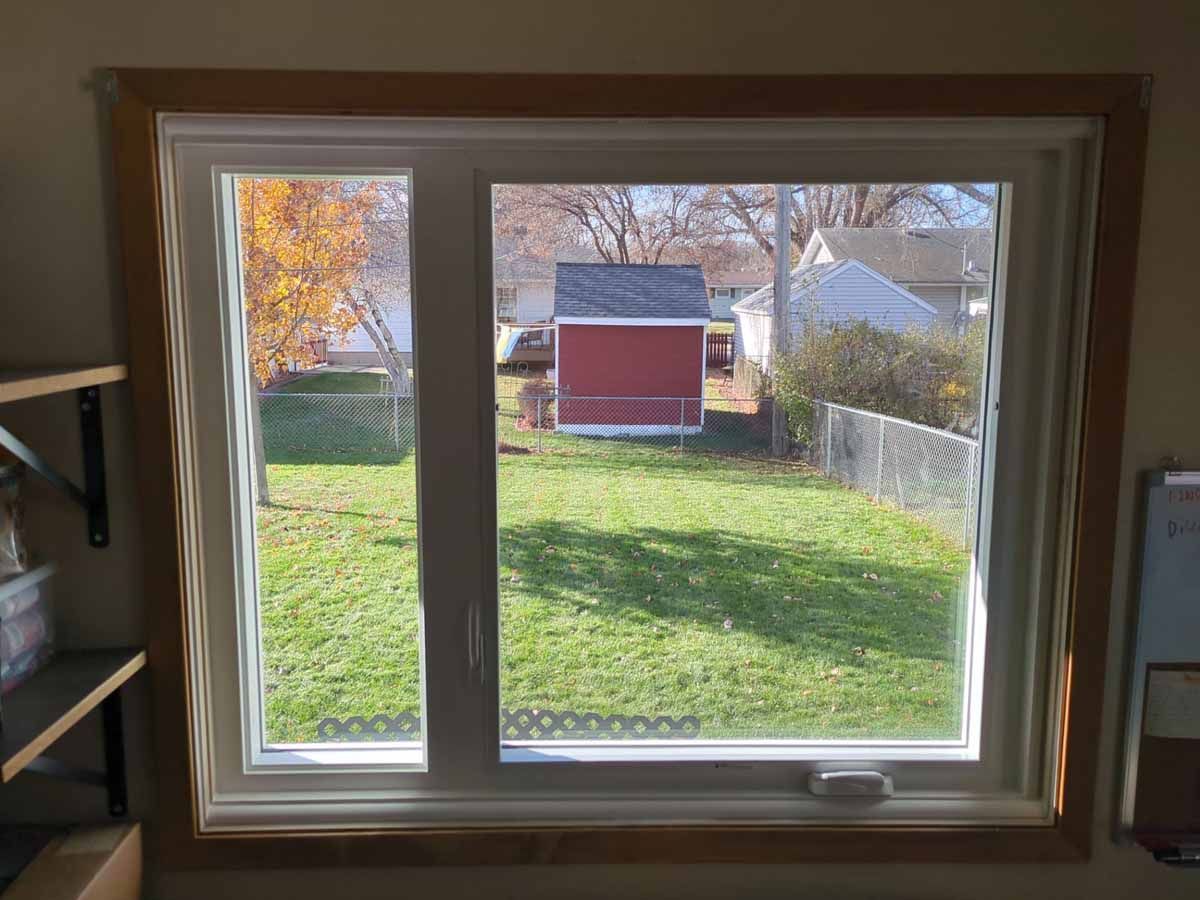 A window framed with wood, looking out onto a backyard with grass, a red shed, and houses.