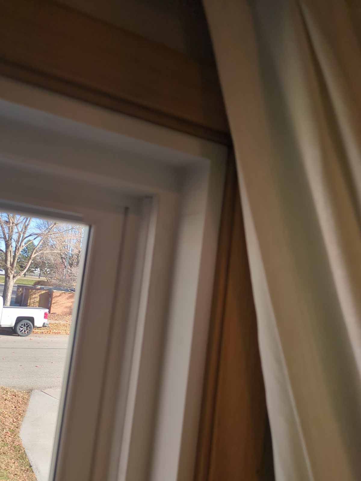 Close-up view of a window with white trim, tan wall, and off-white curtain partially covering the frame; street visible outside.