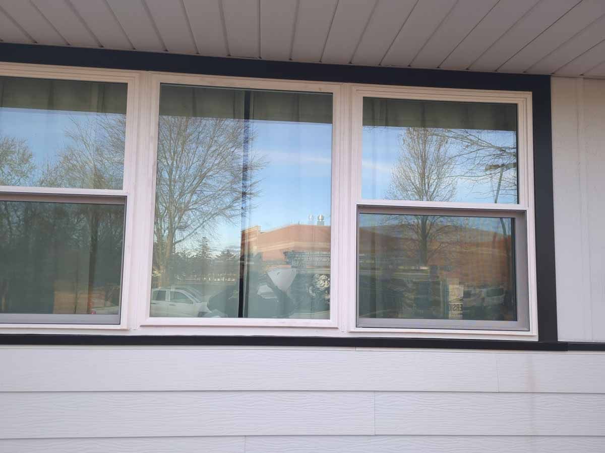 Three white-framed windows with dark trim, reflecting trees and sky against white siding.