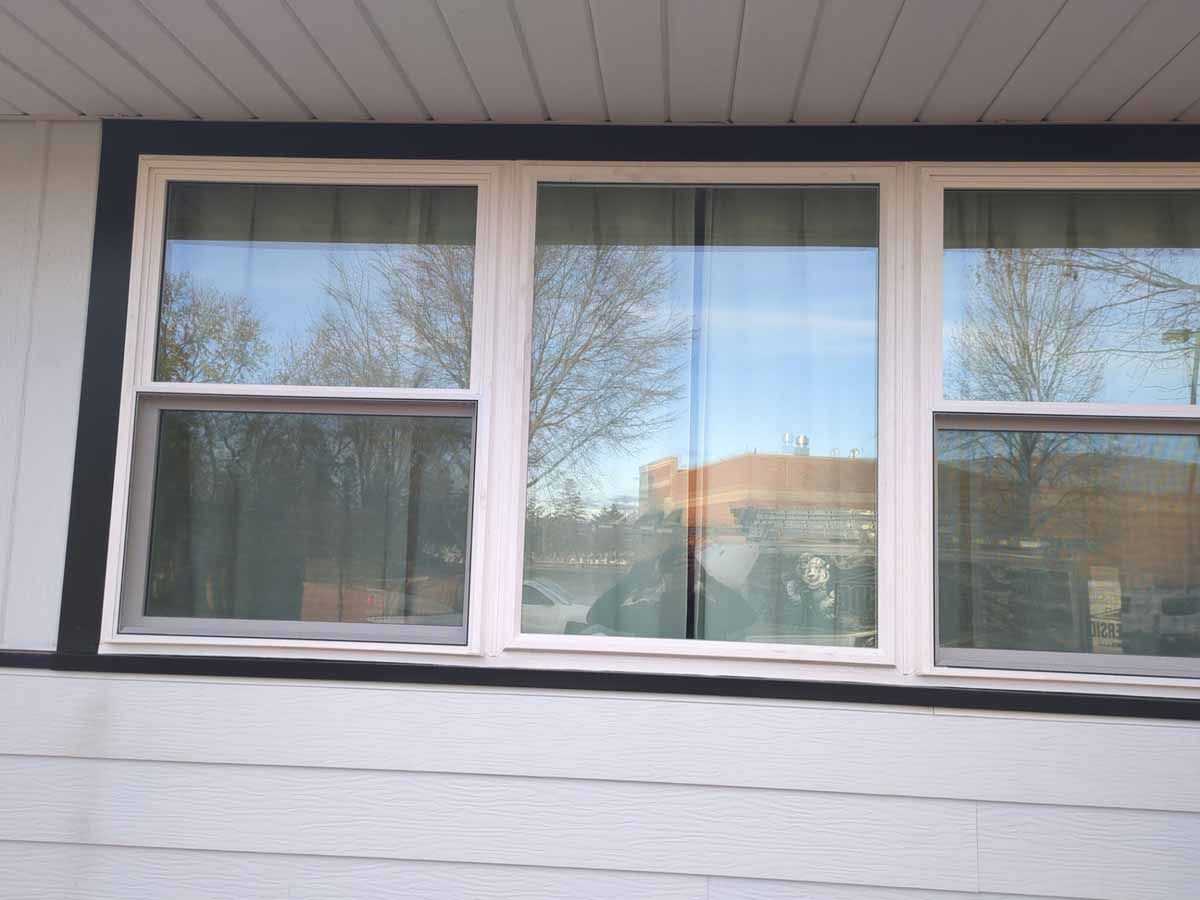 Three-pane window with white frame and dark trim, reflecting blue sky and trees against gray siding.