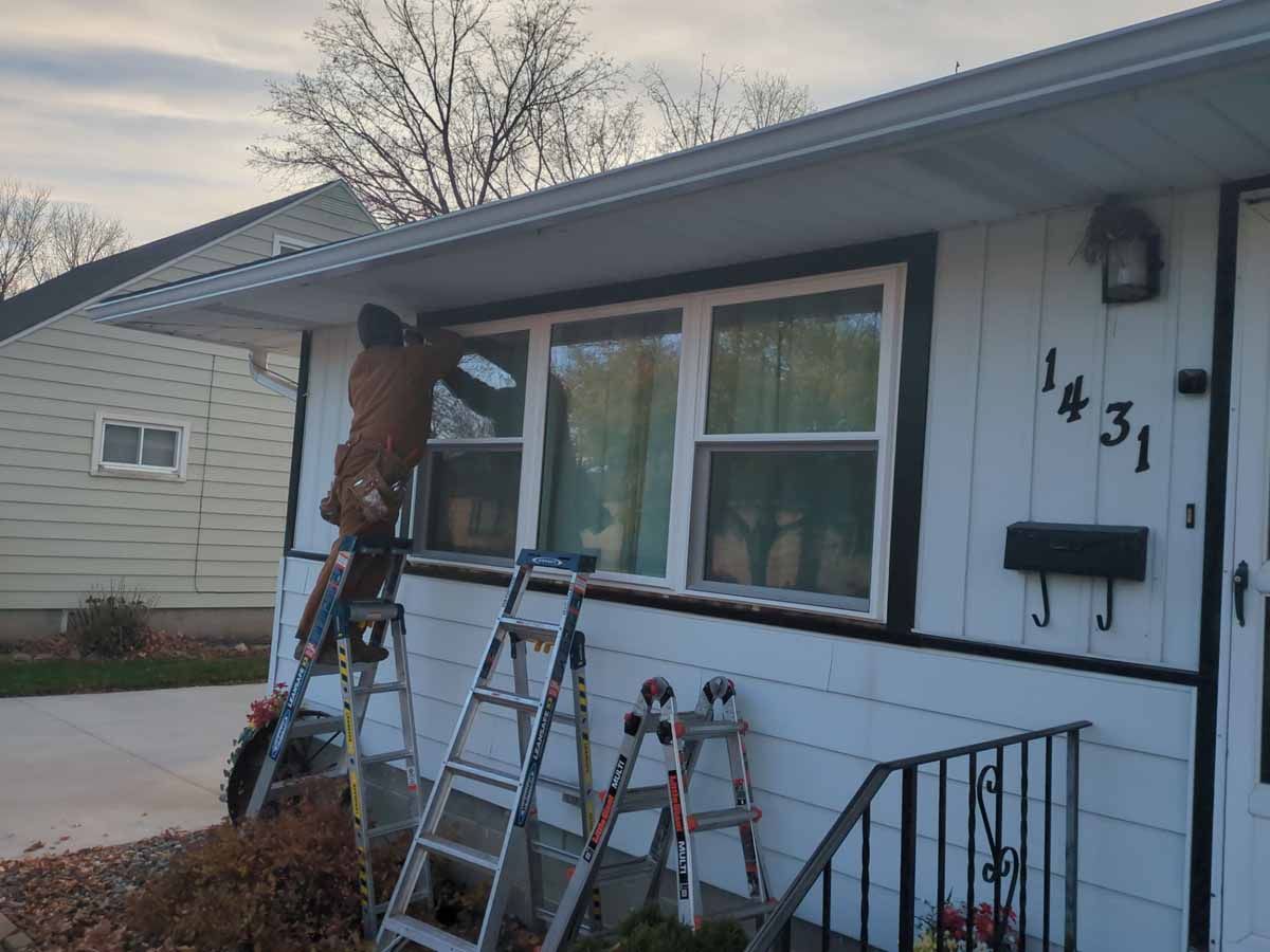 Person on ladder working on the exterior of a white house with black trim. Tools and additional ladder visible.