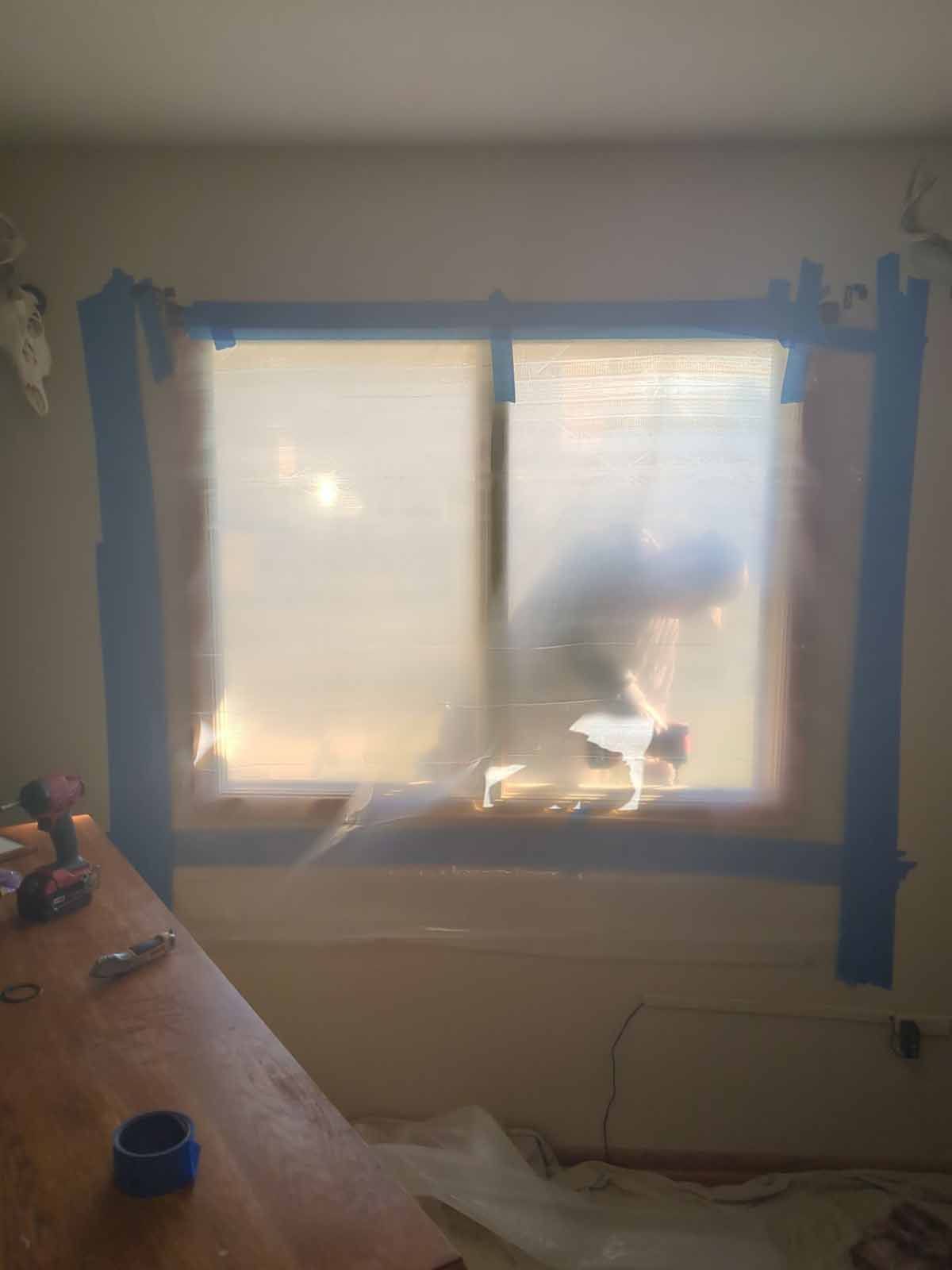 Window covered in plastic for painting, taped around the frame. Work tools are visible on a nearby table.